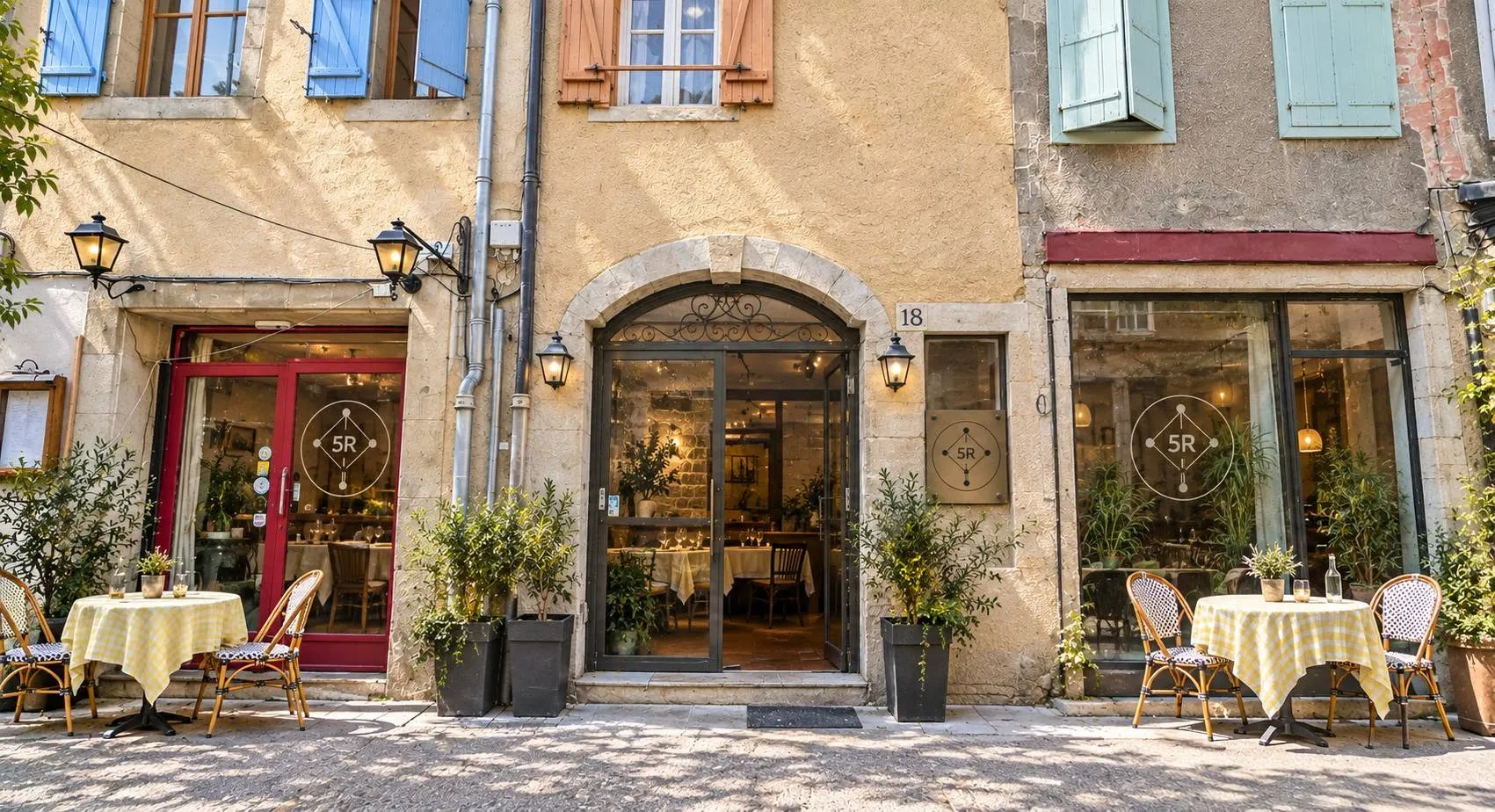 Le 5 R en journée : la porte voûtée du numéro 18 entre des façades de pierre aux volets colorés, et des tables à nappes vichy dressées dans la rue